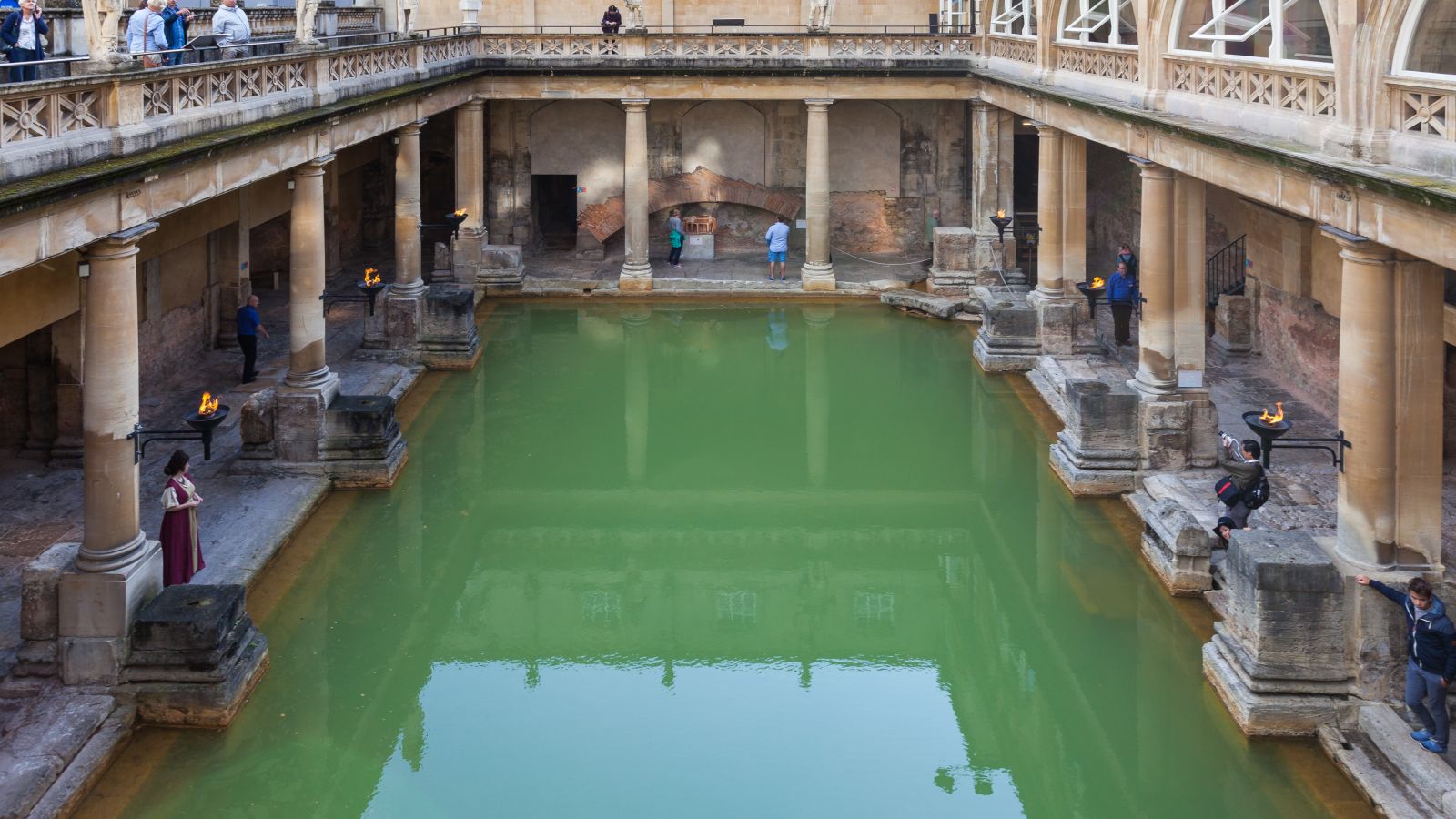 The open-air Great Bath at the Roman Baths in Bath, a rectangular pool of green water edged by weathered stone paving and short columns, with a statue-topped terrace and stone buildings rising behind.