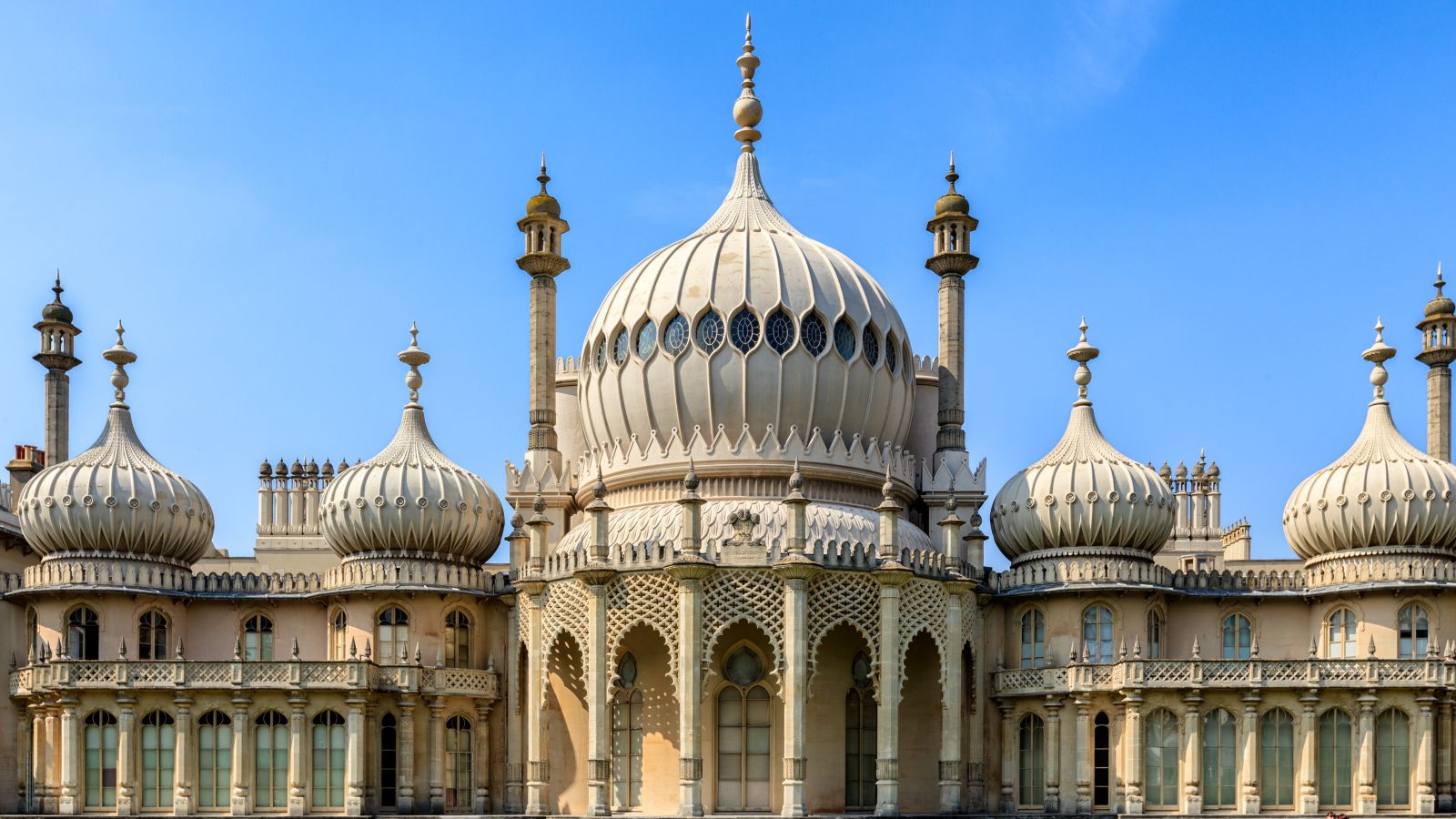 The west front of the Royal Pavilion in Brighton, with a large central onion dome flanked by smaller domes and slender minaret-like towers rising above a colonnade of carved stone arches.