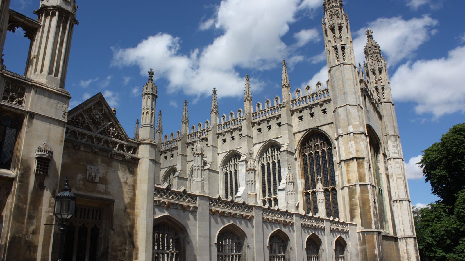 The stone exterior of King's College Chapel in Cambridge seen at an angle from King's Parade, showing its tall traceried windows, buttresses and pinnacled roofline against a blue sky with white clouds.