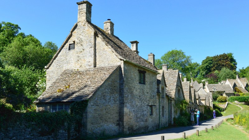 The end cottage of Arlington Row in Bibury, a steep-gabled building of pale Cotswold stone with a stone-tiled roof and two chimneys, with the rest of the terrace and a narrow lane receding behind it.