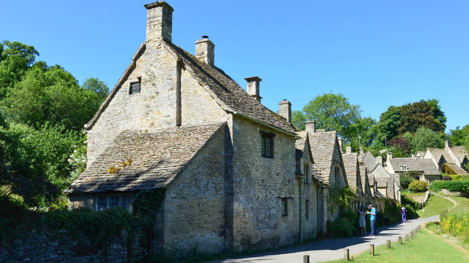 The end cottage of Arlington Row in Bibury, a steep-gabled building of pale Cotswold stone with a stone-tiled roof and two chimneys, with the rest of the terrace and a narrow lane receding behind it.