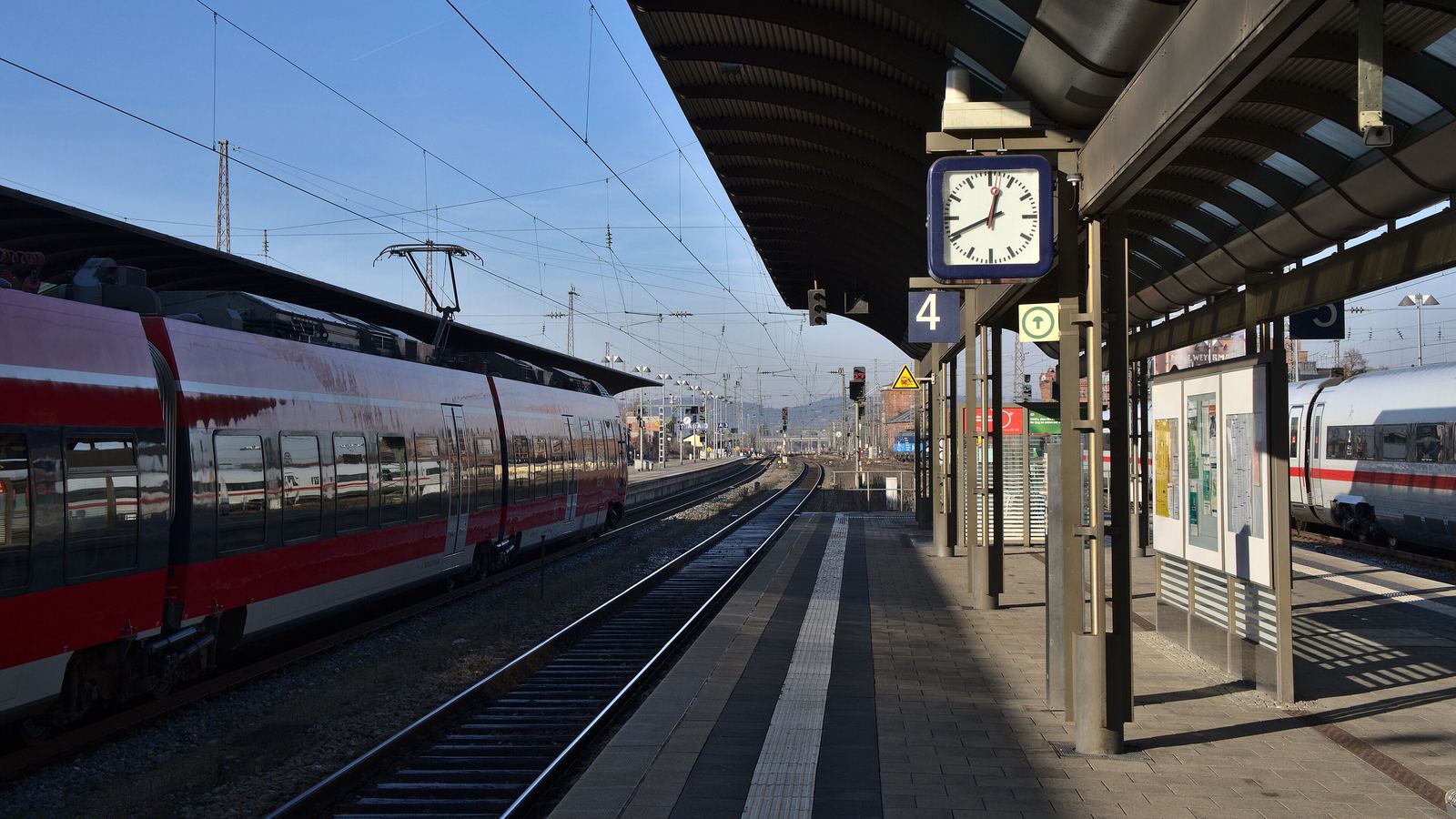 A sunlit station platform at Bamberg, with a red and white regional train standing at the track and a round platform clock hanging under the canopy.