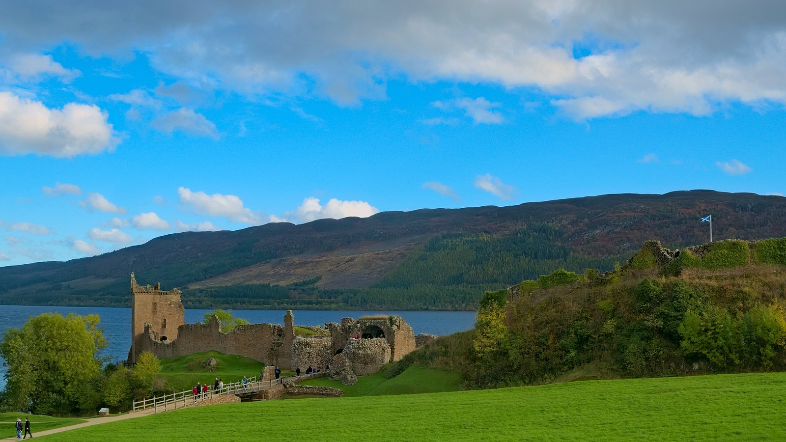 The stone ruins of Urquhart Castle on a grassy headland beside Loch Ness, with hills along the far shore under a blue sky.