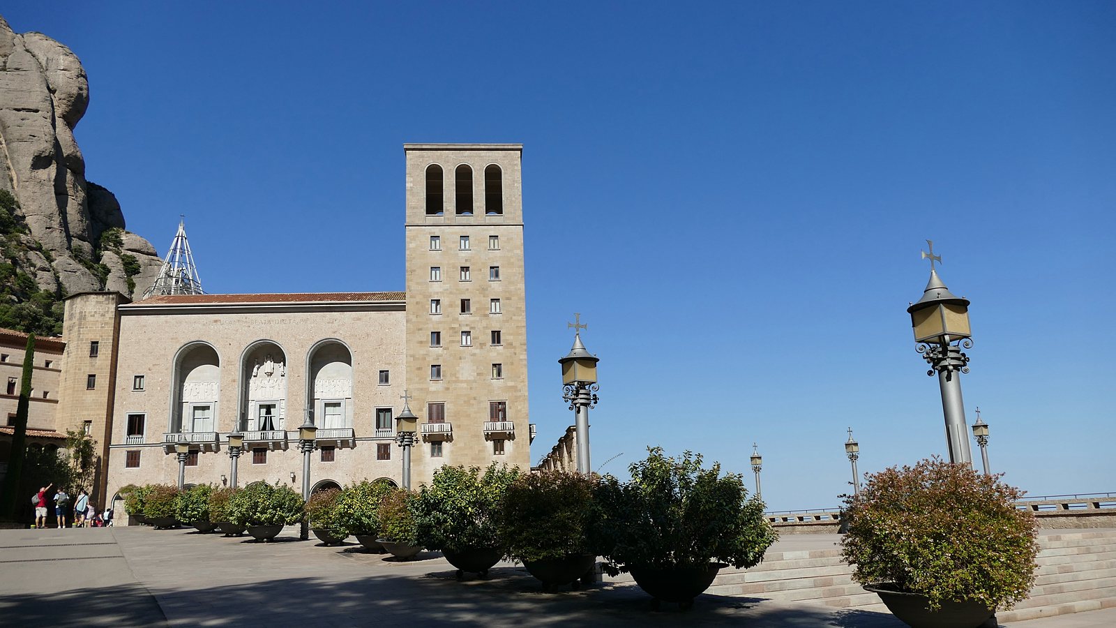 The stone front and square bell tower of Montserrat monastery seen across the paved Plaça de Santa Maria, with the serrated rock of the mountain rising at the left.