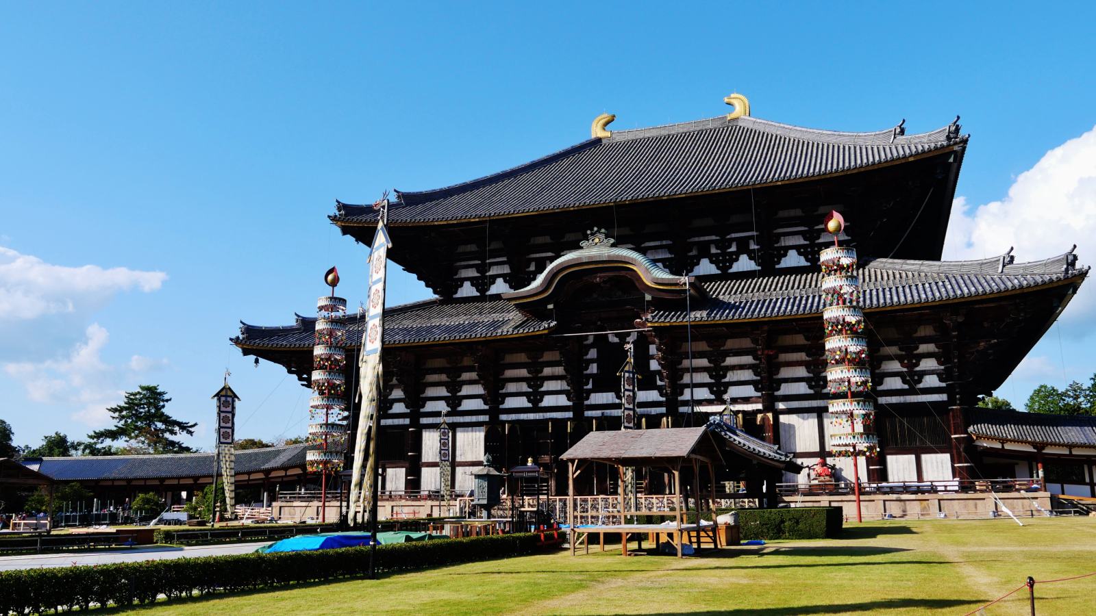 The large timber Daibutsuden hall of Todai-ji in Nara viewed from the south across a mown lawn, with two gilded ornaments on its tiled roof ridge and a tall banner pole standing on each side.