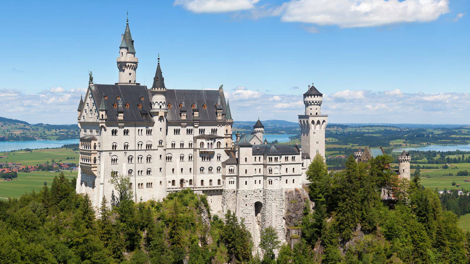 Neuschwanstein Castle, pale stone with grey turrets and a tall keep, standing on a wooded hill above a green valley and a lake.