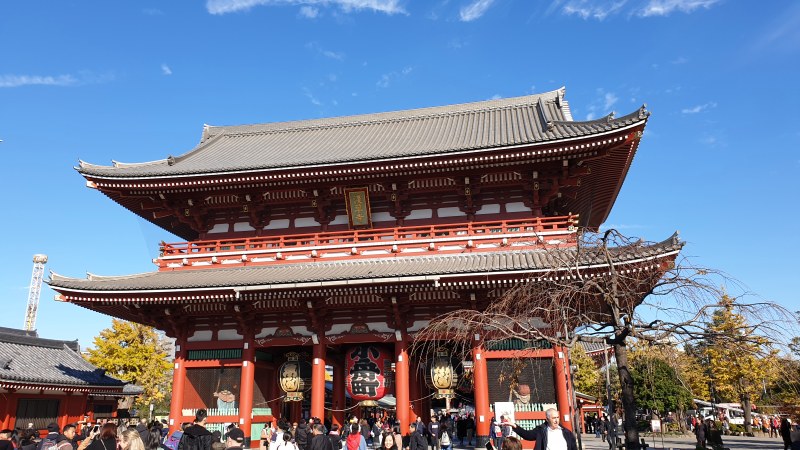 The two-storey Hozomon gate at Senso-ji temple in Asakusa, Tokyo, seen from below against a clear blue sky, with red-painted timber columns, two tiled roofs and large paper lanterns hanging in its openings.