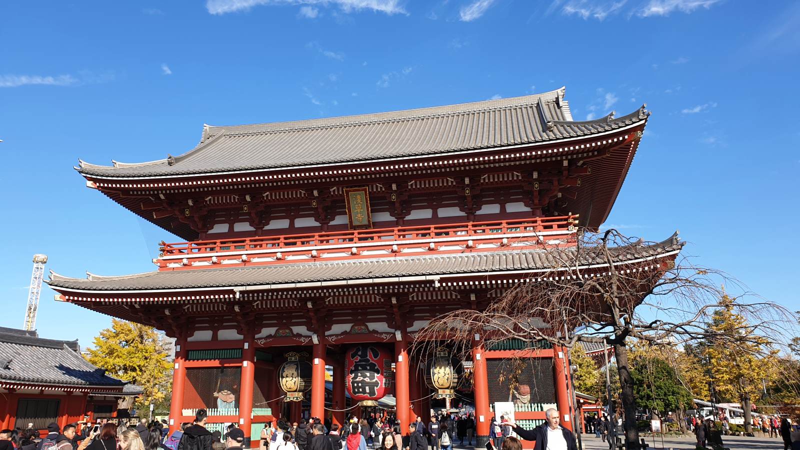 The two-storey Hozomon gate at Senso-ji temple in Asakusa, Tokyo, seen from below against a clear blue sky, with red-painted timber columns, two tiled roofs and large paper lanterns hanging in its openings.