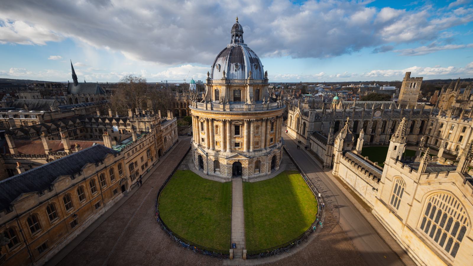 An elevated view over Radcliffe Square in Oxford, with the domed circular Radcliffe Camera standing on a lawn enclosed on all sides by stone college and library buildings, and city rooftops beyond.