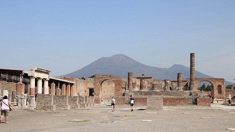 The open forum of Pompeii, lined with broken columns and brick walls, with the cone of Mount Vesuvius rising behind it.