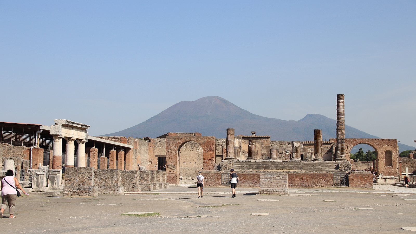 The open forum of Pompeii, lined with broken columns and brick walls, with the cone of Mount Vesuvius rising behind it.