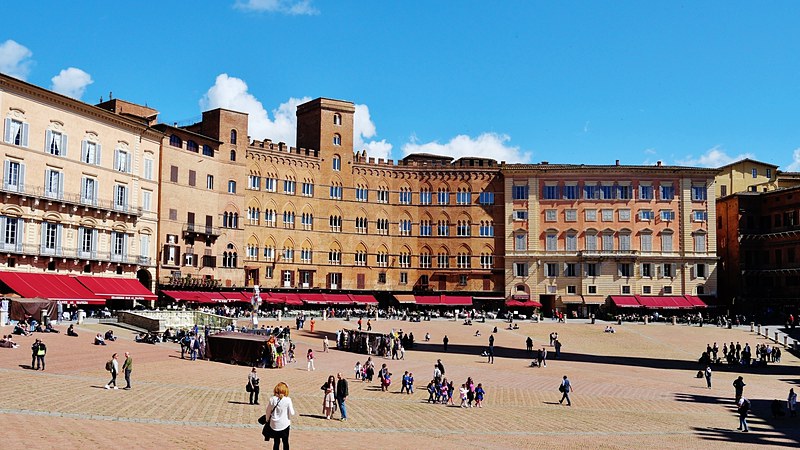Brick and stone palaces lining the curve of the Piazza del Campo in Siena, with red cafe awnings at street level and people crossing the sloping square.