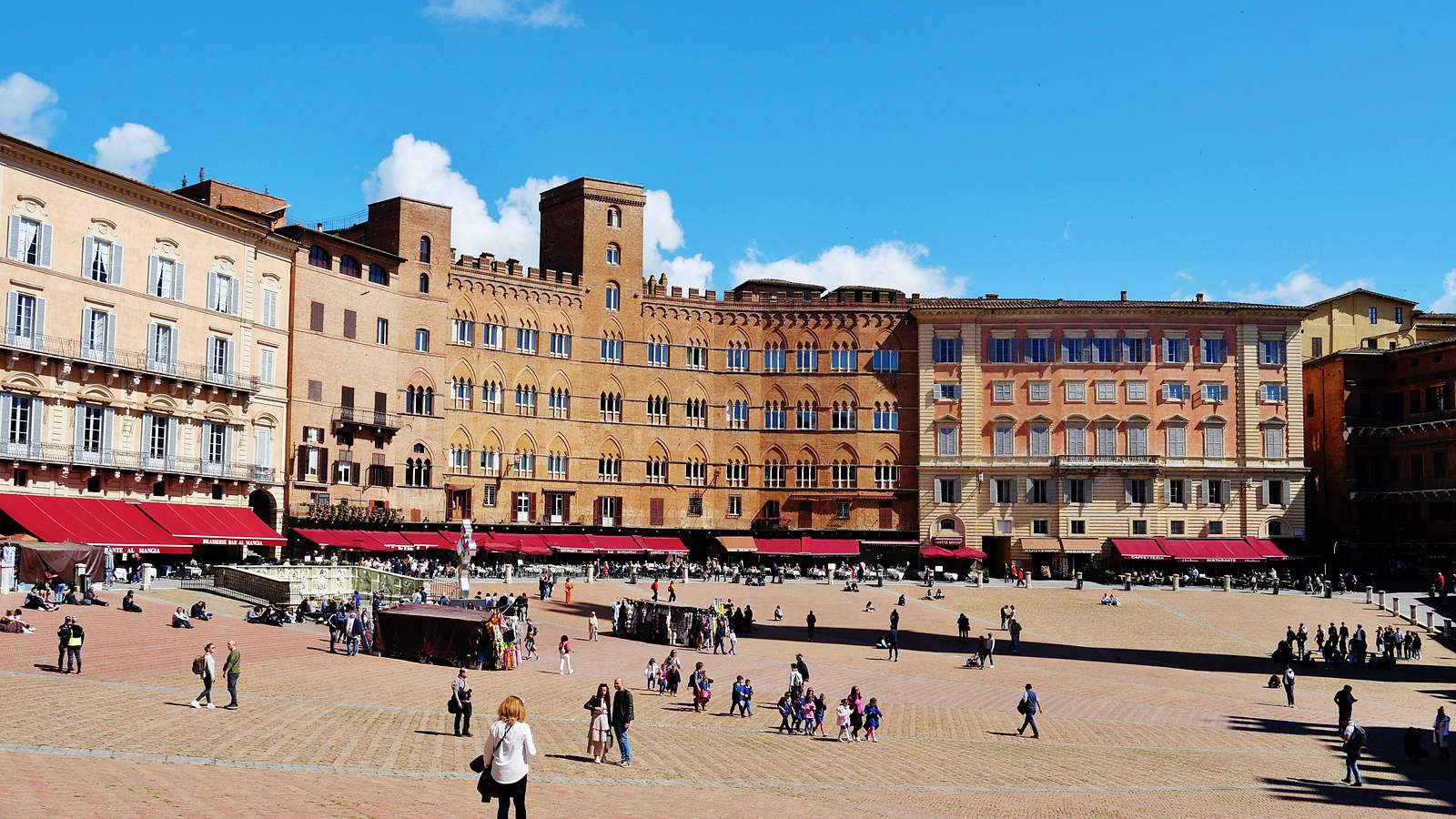 Brick and stone palaces lining the curve of the Piazza del Campo in Siena, with red cafe awnings at street level and people crossing the sloping square.