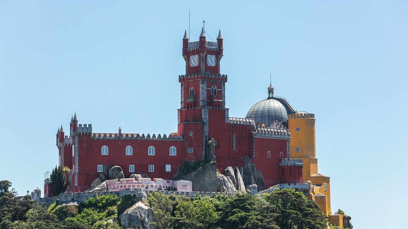 The Pena Palace on its wooded hilltop in Sintra, with a red clock tower, a grey dome and a yellow wing, under a clear blue sky.