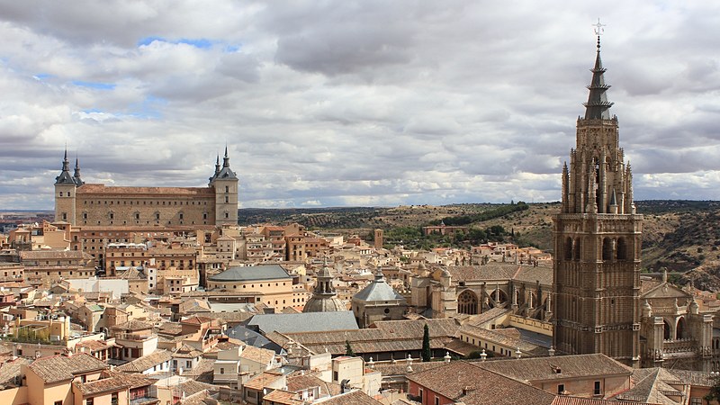 A view across the tiled rooftops of Toledo, with the square Alcázar fortress on the left and the spired bell tower of the cathedral on the right under a cloudy sky.