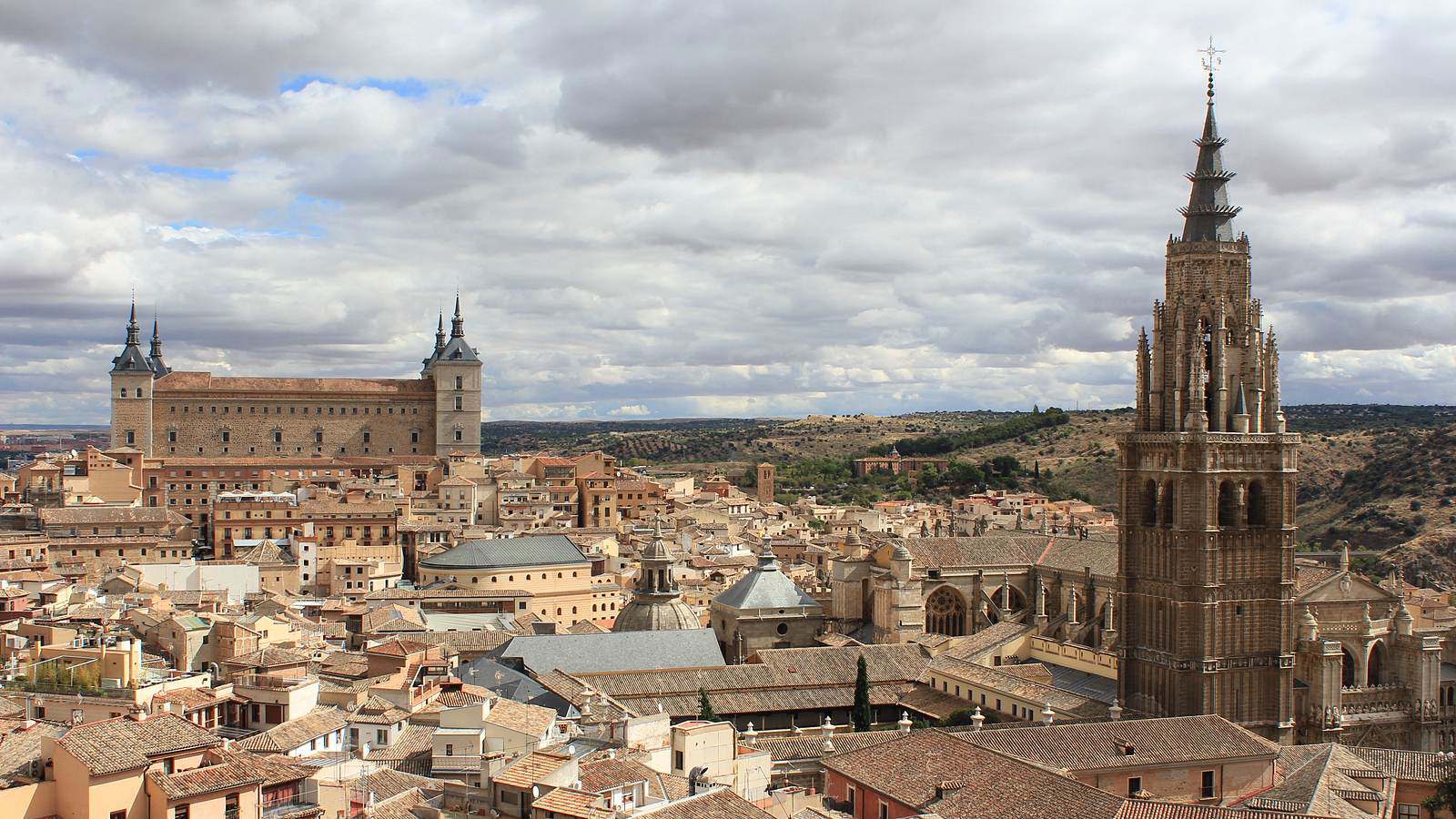 A view across the tiled rooftops of Toledo, with the square Alcázar fortress on the left and the spired bell tower of the cathedral on the right under a cloudy sky.