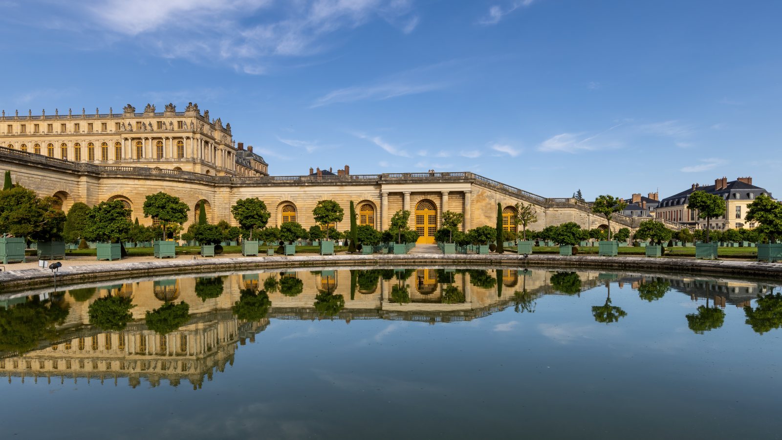 The long arcaded Orangerie building at the Palace of Versailles, fronted by rows of clipped trees in square planters and reflected in the still water of a rectangular pond under a blue sky.