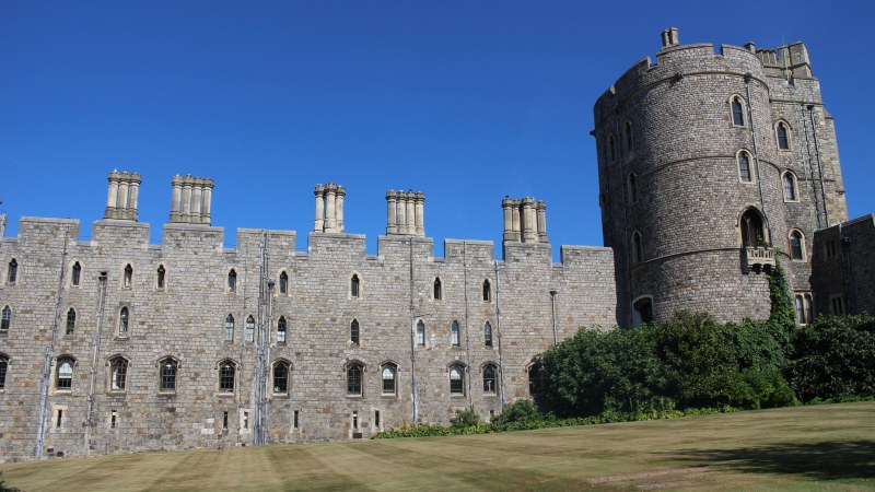 The Round Tower of Windsor Castle standing on its mound at the right, with the battlemented stone range of the Upper Ward and its row of chimneys extending to the left above a lawn.
