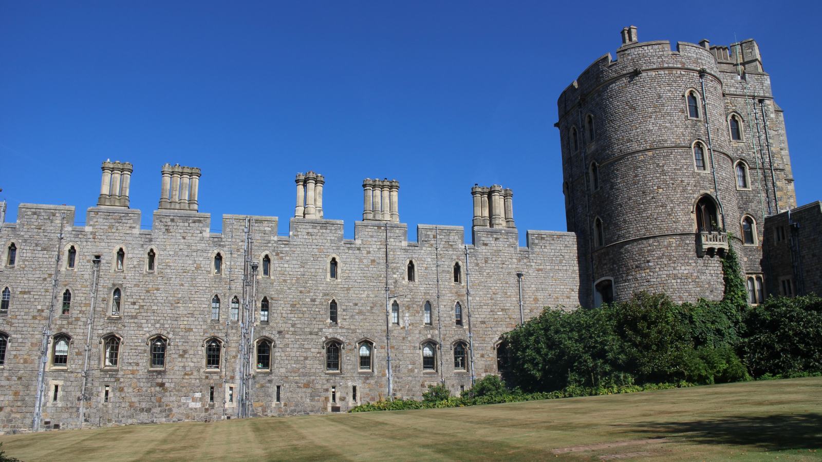 The Round Tower of Windsor Castle standing on its mound at the right, with the battlemented stone range of the Upper Ward and its row of chimneys extending to the left above a lawn.